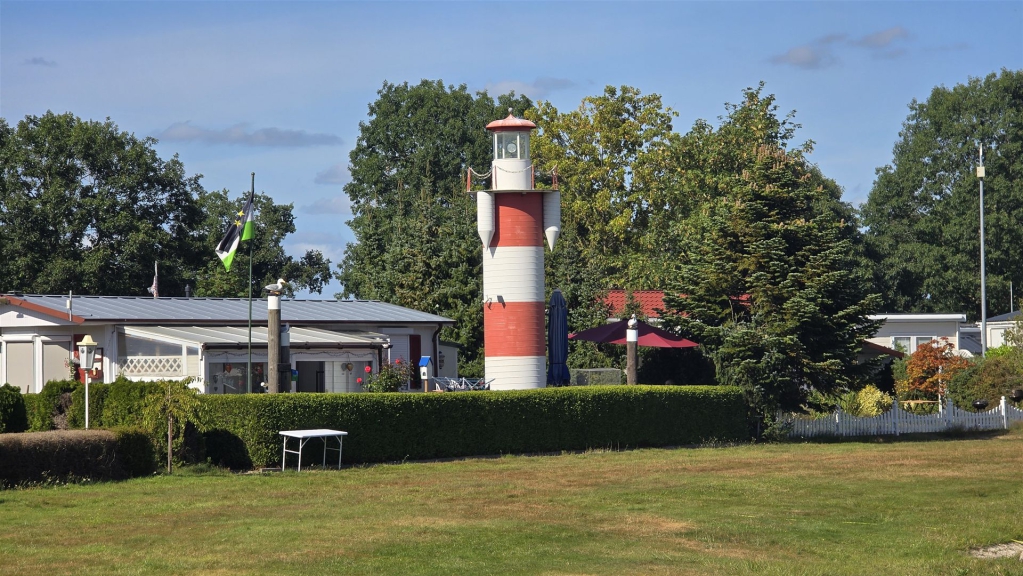 Ein rot-weiß gestreifter Leuchtturm inmitten eines Parks mit viel Grün und Gebäuden im Hintergrund.