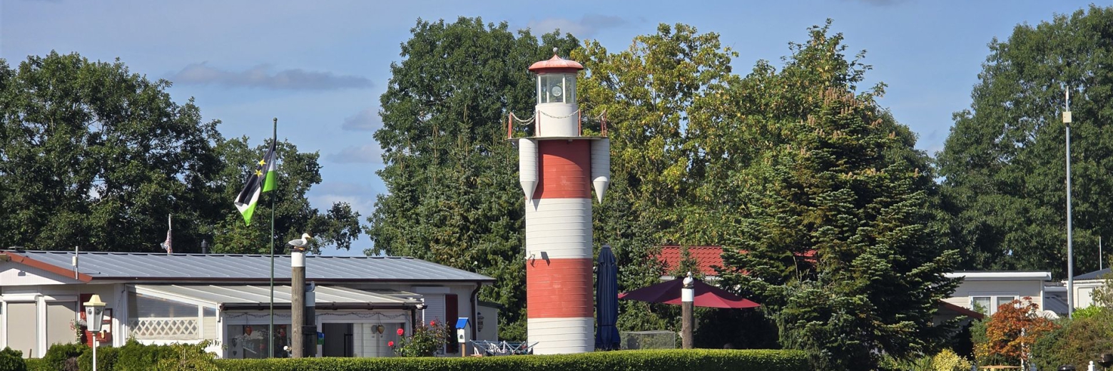 Ein markanter Leuchtturm mit roten und weißen Streifen steht vor einer Kulisse aus Bäumen, Gebäuden und einem gepflegten Rasen.