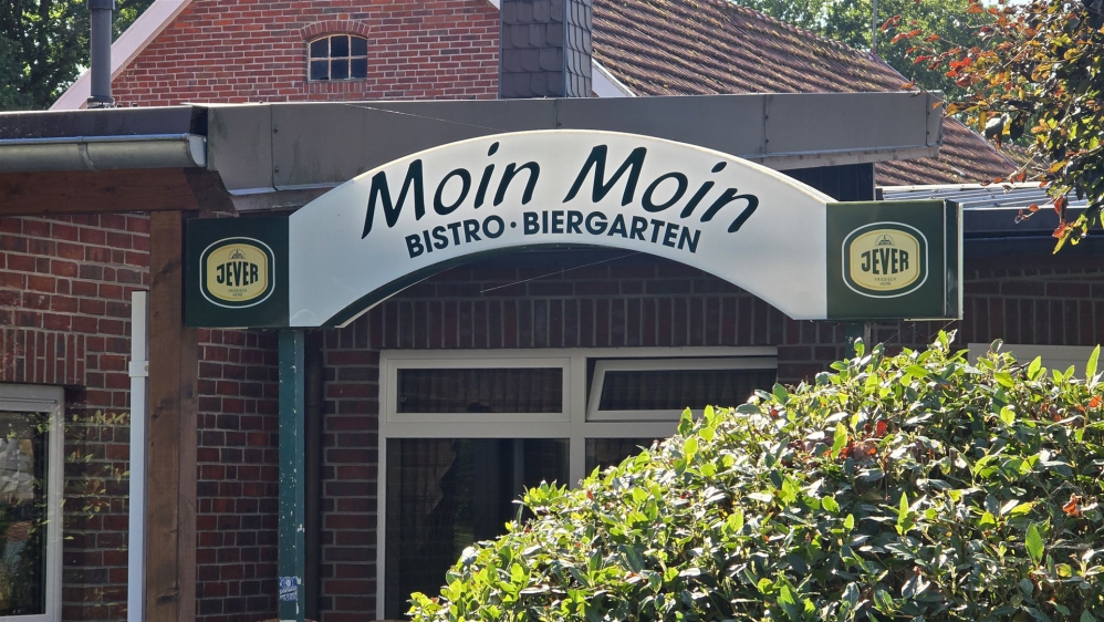 Fassade eines Restaurants mit dem Schild Moin Moin Bistro Biergarten, teilweise verdeckt von Grünpflanzen.