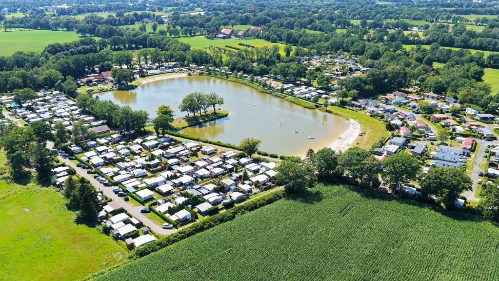 Luftbild eines großen Campingplatzes, der einen zentralen See umgibt und von Feldern sowie Waldgebieten gesäumt wird.