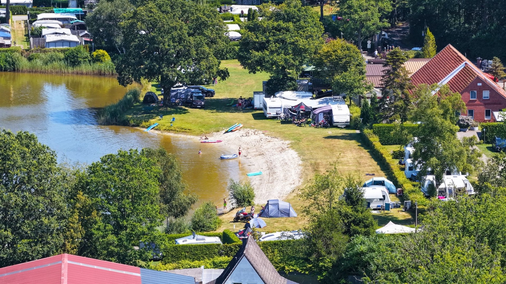 Weitwinkelaufnahme eines Campingplatzes an einem großen See mit Wäldern und Gebäuden im Hintergrund.